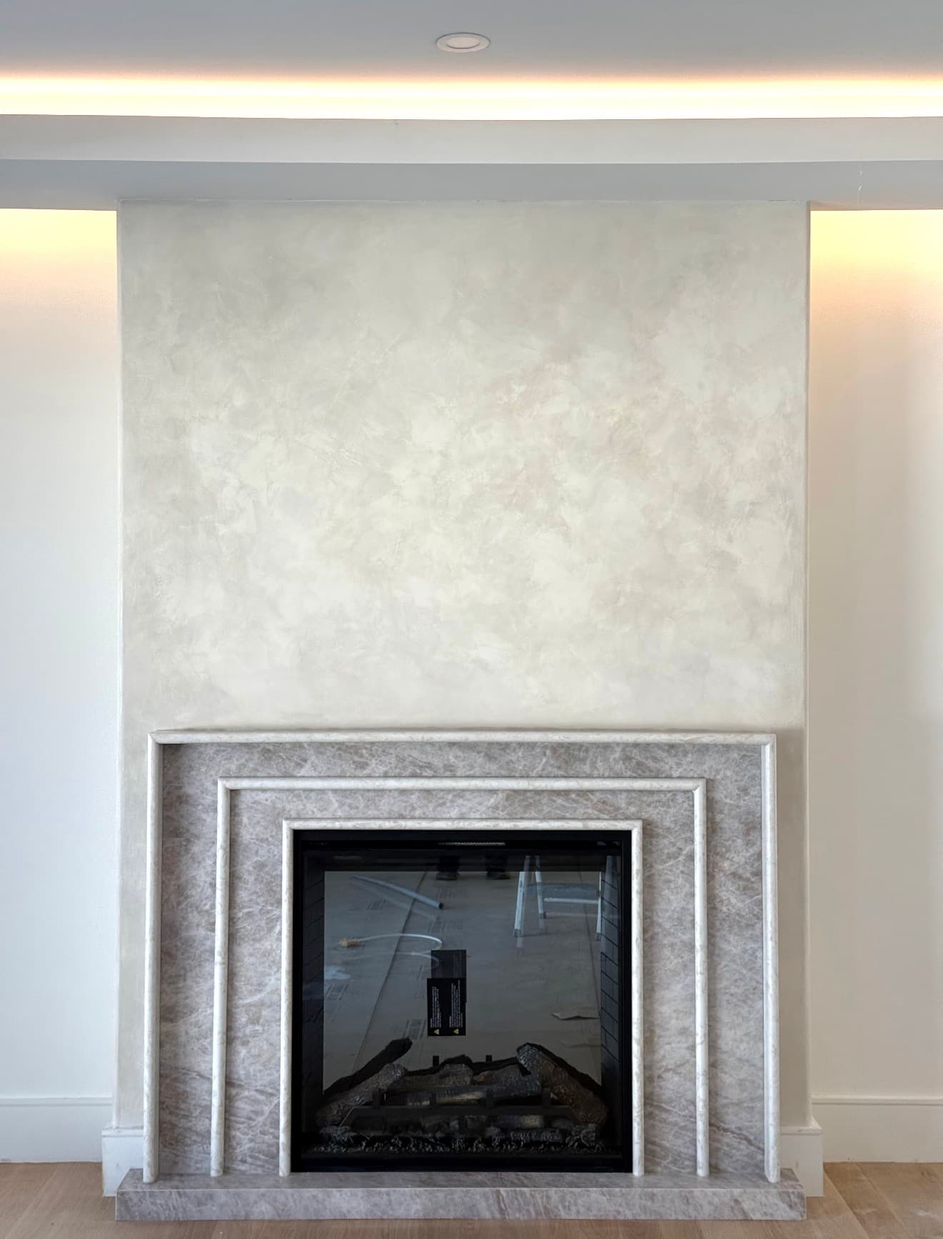 A fireplace with a pale plaster chimney breast above a stone surround, lit from a cove in the ceiling above.