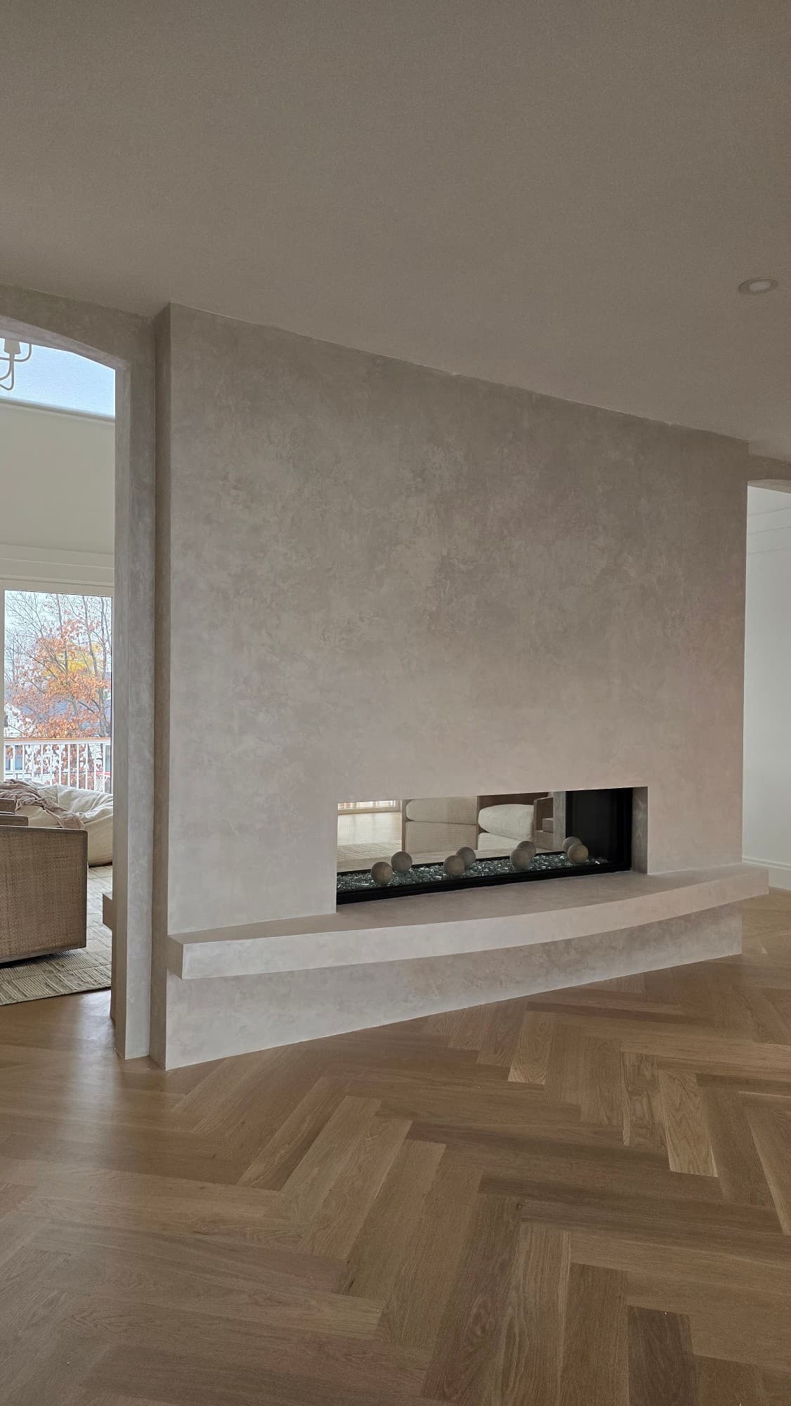 A full-height plaster feature wall in a living room, wrapping a linear fireplace with a curved built-in hearth, over a herringbone wood floor.