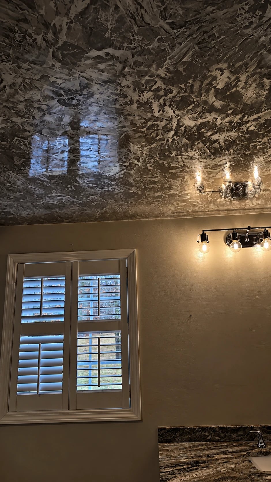 A bathroom ceiling in dark Grassello Fino, polished to a high gloss that reflects the window and the vanity lights below.