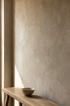 Low sunlight raking across a pale lime-washed wall above a timber bench and a ceramic bowl.