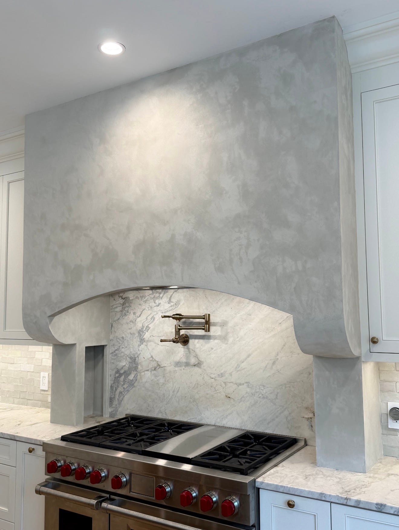 A kitchen range hood finished in pale grey Marmorino, its curved skirt running over a marble backsplash and a professional range.