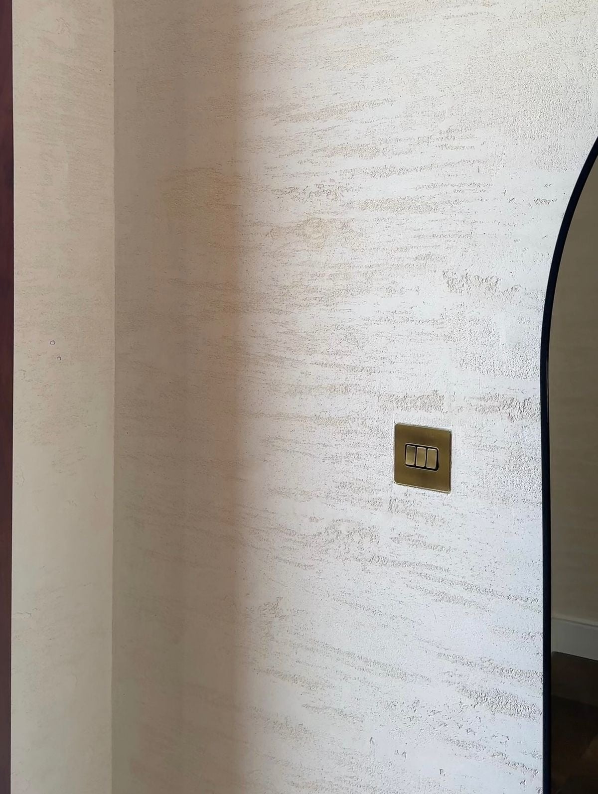 Close view of the travertine-effect finish: horizontal drag marks and open pores in a warm off-white surface, beside a brass switch plate and an arched mirror.