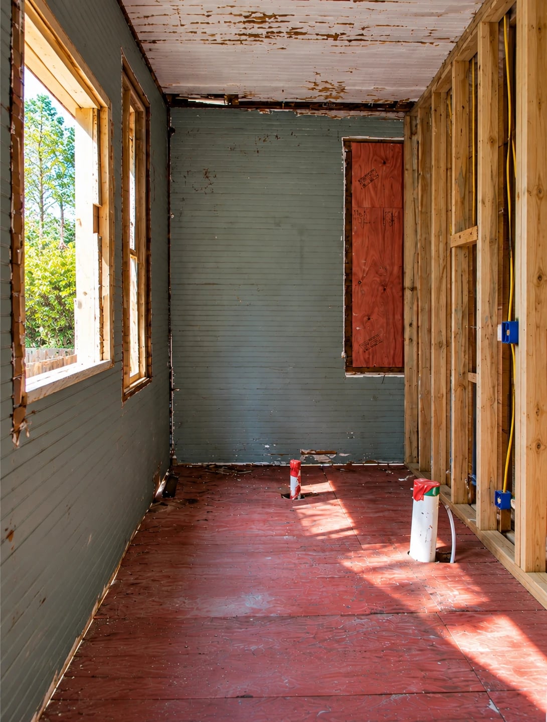 A gutted room down to the studs: open timber framing and new wiring on one side, painted board on the others, and plumbing stubs rising from a bare subfloor.
